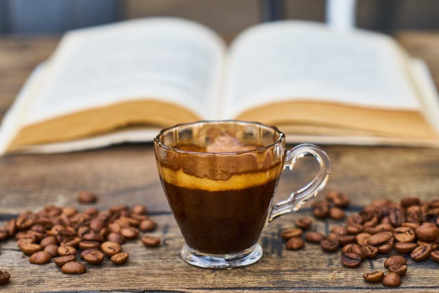 A glass of coffee in front of a book, surrounded by coffee beans.